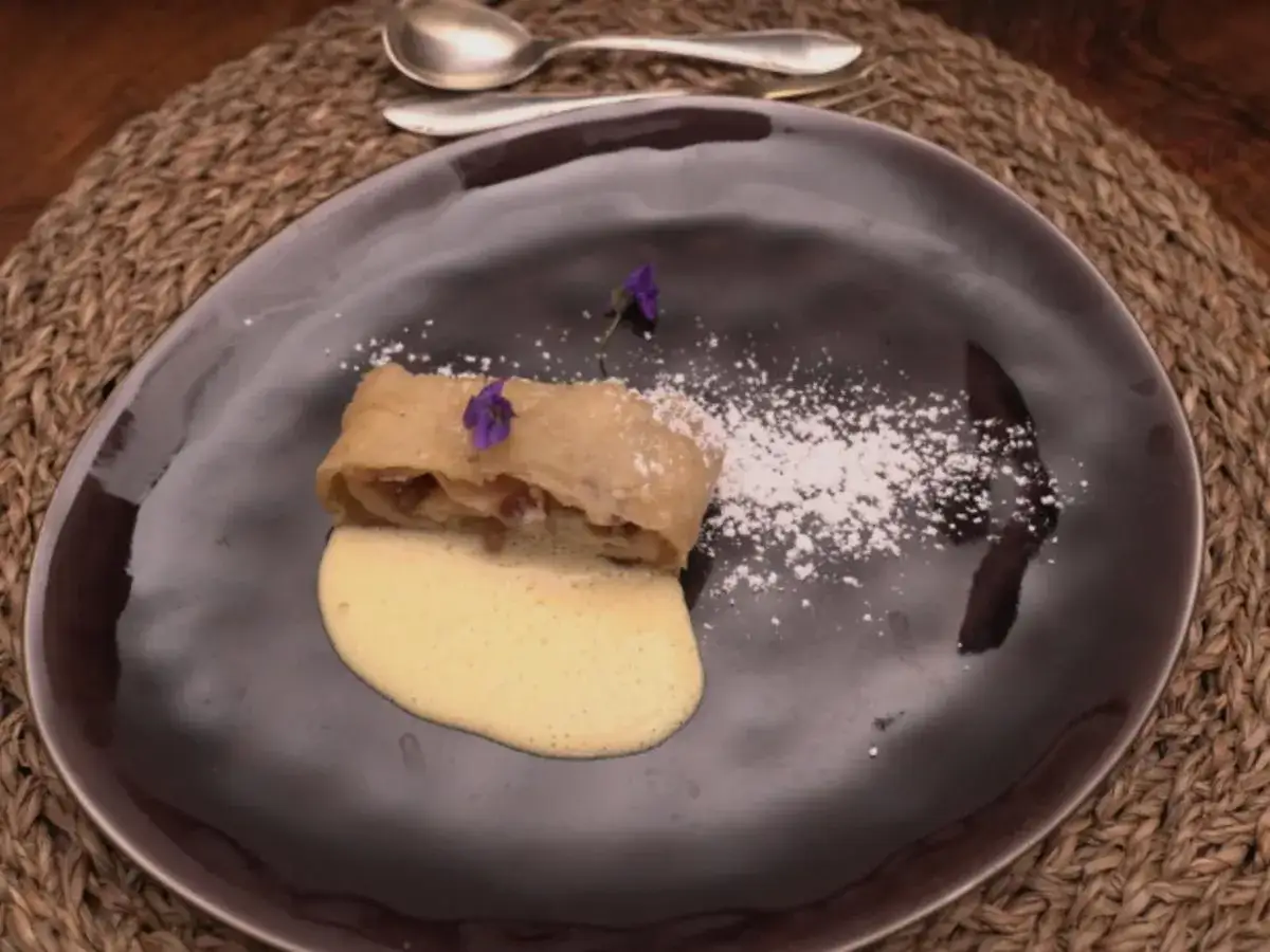 Ein St&uuml;ck bayerischer Apfelstrudel mit Vanillesauce und Puderzucker bestreut, garniert mit einer kleinen lila Bl&uuml;te.