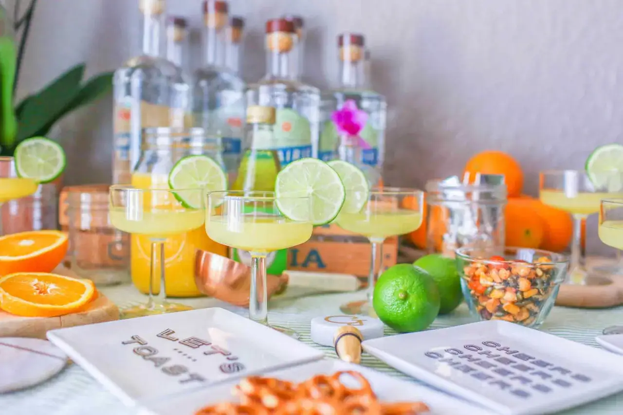 Gotowy drink z ginem, ozdobiony plasterkiem limonki, czeka na toast. Na stole butelki ginu, pomarańcze i przekąski.