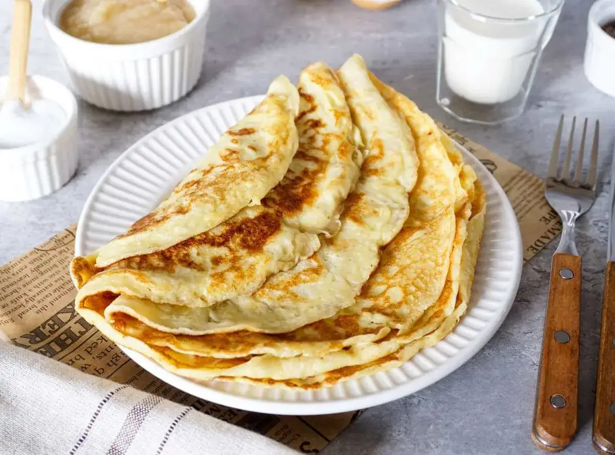 Ein Stapel goldbrauner Pfannkuchen, perfekt f&uuml;r ein sehr gutes Pfannkuchen Rezept. Dazu Apfelmus, Zucker und Milch.