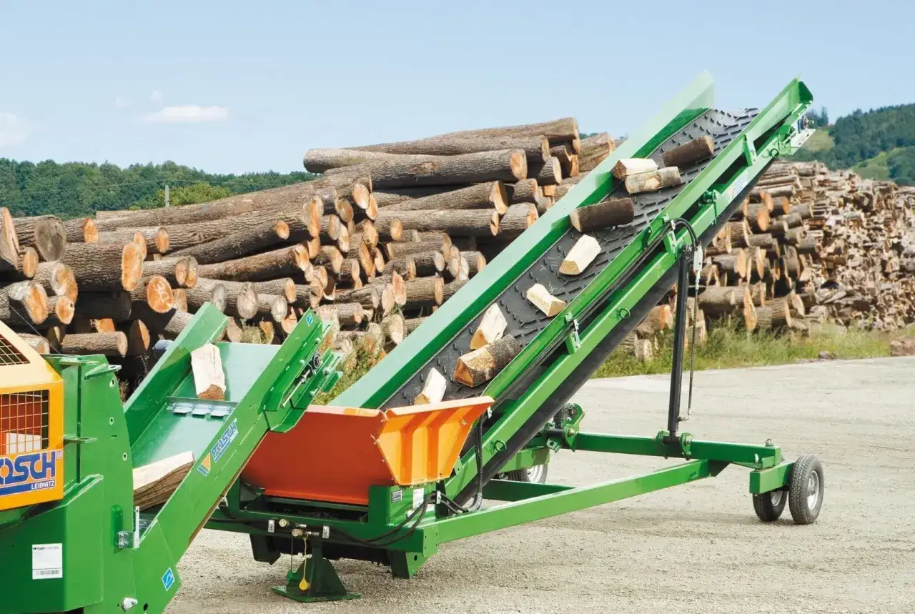 Gr&uuml;ne mobile F&ouml;rderb&auml;nder transportieren Holzscheite vor einem gro&szlig;en Stapel Baumst&auml;mme.