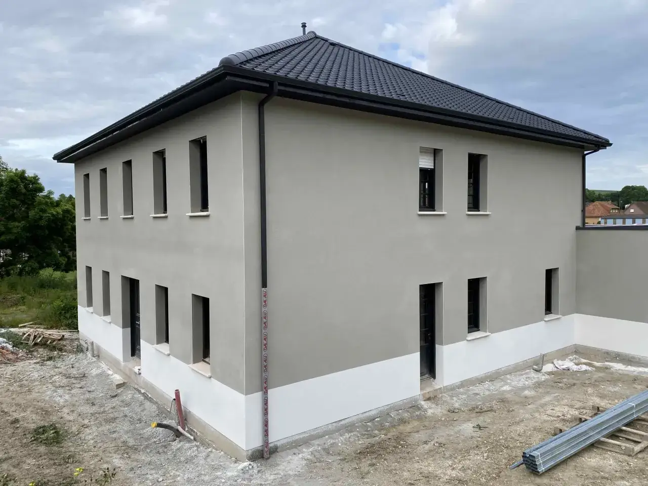 Maison moderne avec un enduit mur extérieur gris clair et une bande blanche à la base. Le toit est couvert de tuiles sombres.