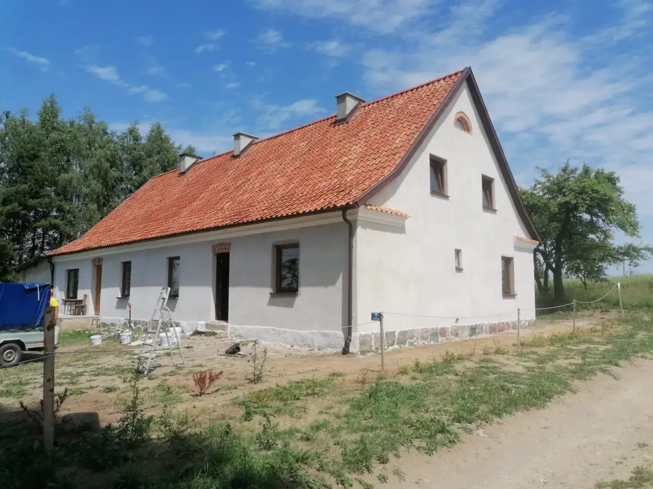 Dom z czerwonym dachem, otynkowany, z kamiennym cokołem. Widać drabiny i wiadra, sugerujące prace wykończeniowe z użyciem wapna gaszonego.