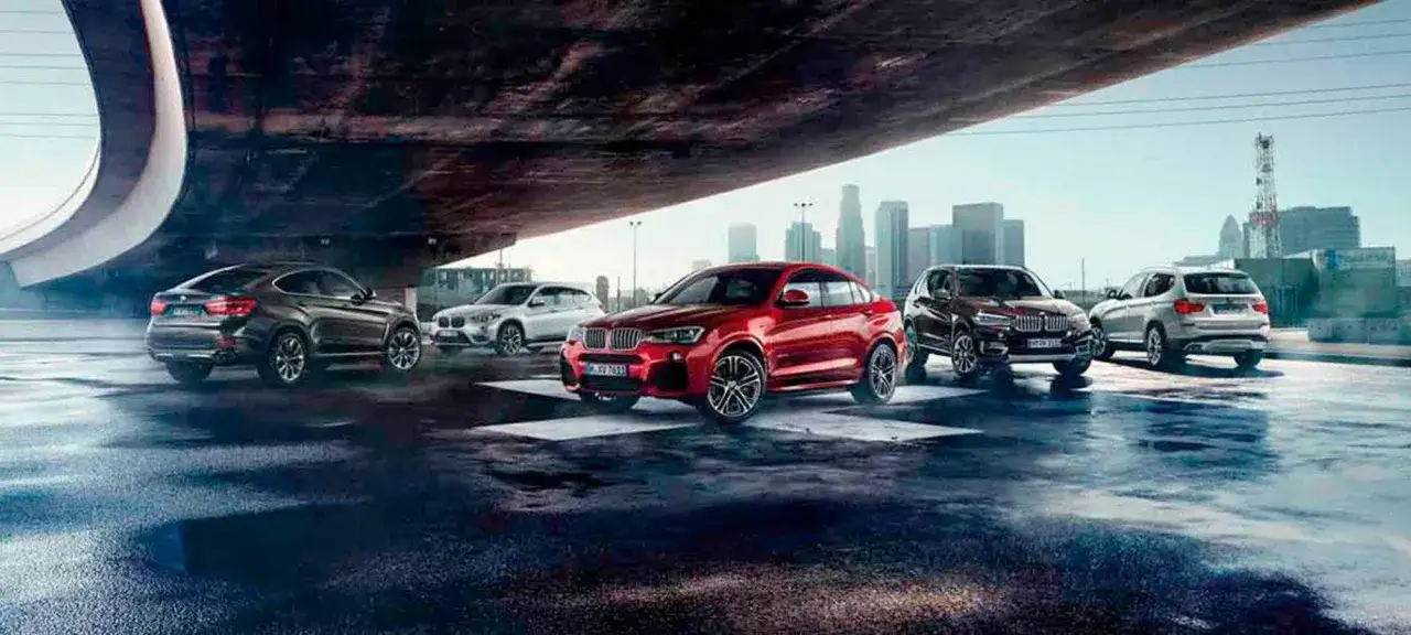 Cinco veh&iacute;culos de la bmw serie x, incluyendo un rojo deportivo, se alinean bajo un puente con una ciudad al fondo.