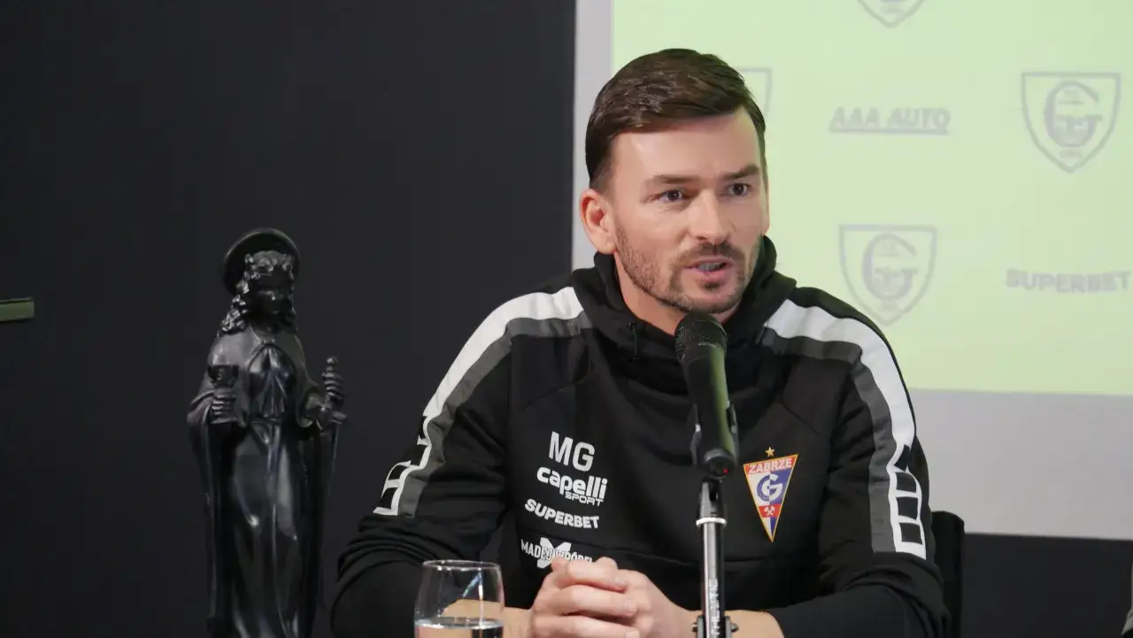 Trener Górnika Zabrze w czarnej bluzie z logo klubu, siedzi przy mikrofonie obok figury świętego.