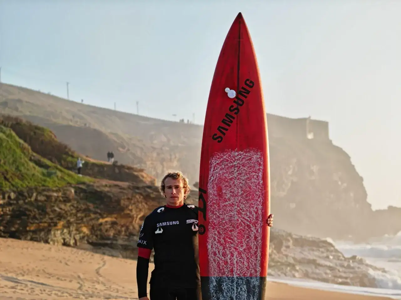 Surfer z czerwoną deską Samsung, gotowy na fale. Czeka na idealny moment, by zanurkować pod falę.