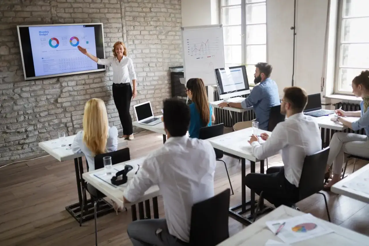 A presenter explains a monthly budget chart on a screen to an audience in a modern classroom.