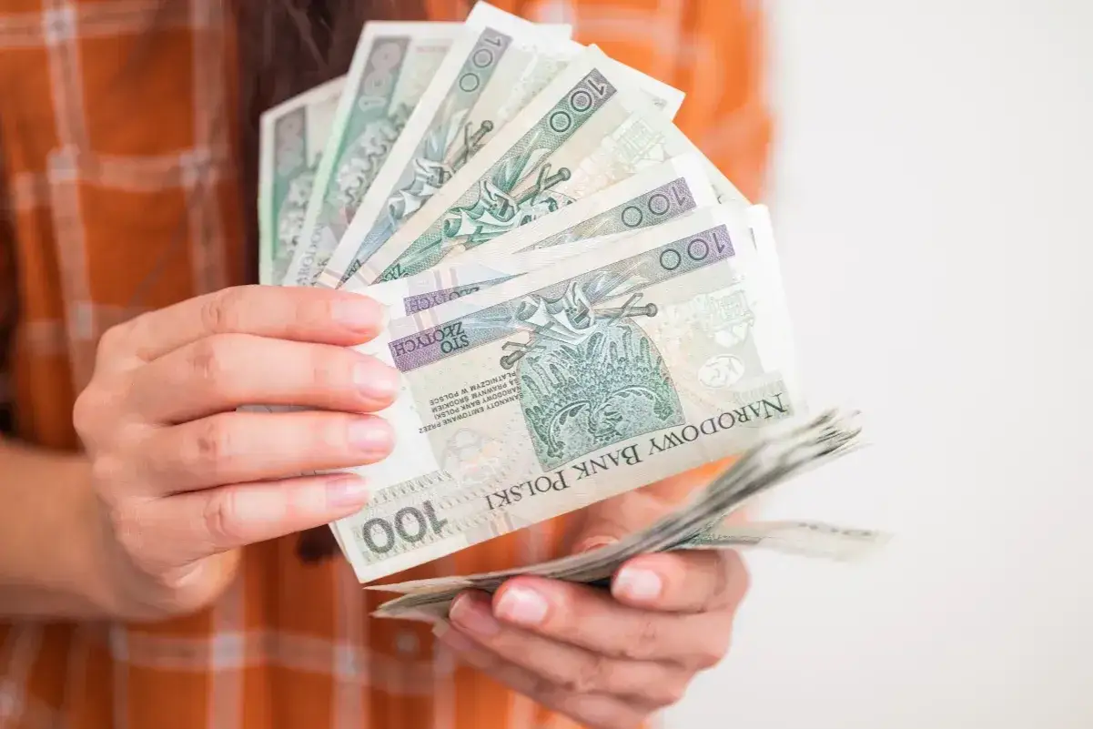 Dłonie trzymające stos banknotów 100 zł. Zastanawiasz się, ile wynosi zasiłek dla bezrobotnych?