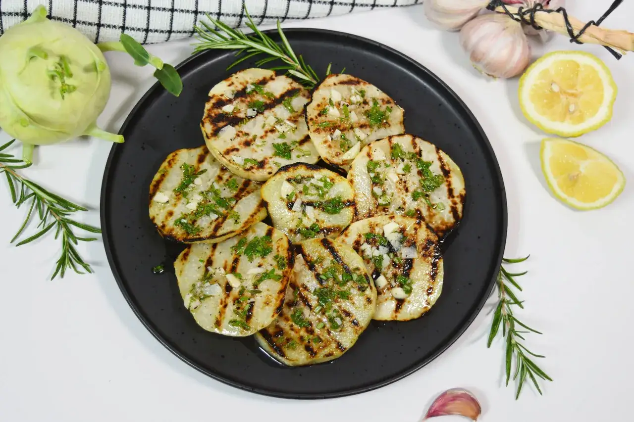 Gegrillte Kohlrabi-Scheiben mit Kräutern und Knoblauch auf schwarzer Platte, garniert mit Rosmarin. Dazu Zitronenspalten und Knoblauch.