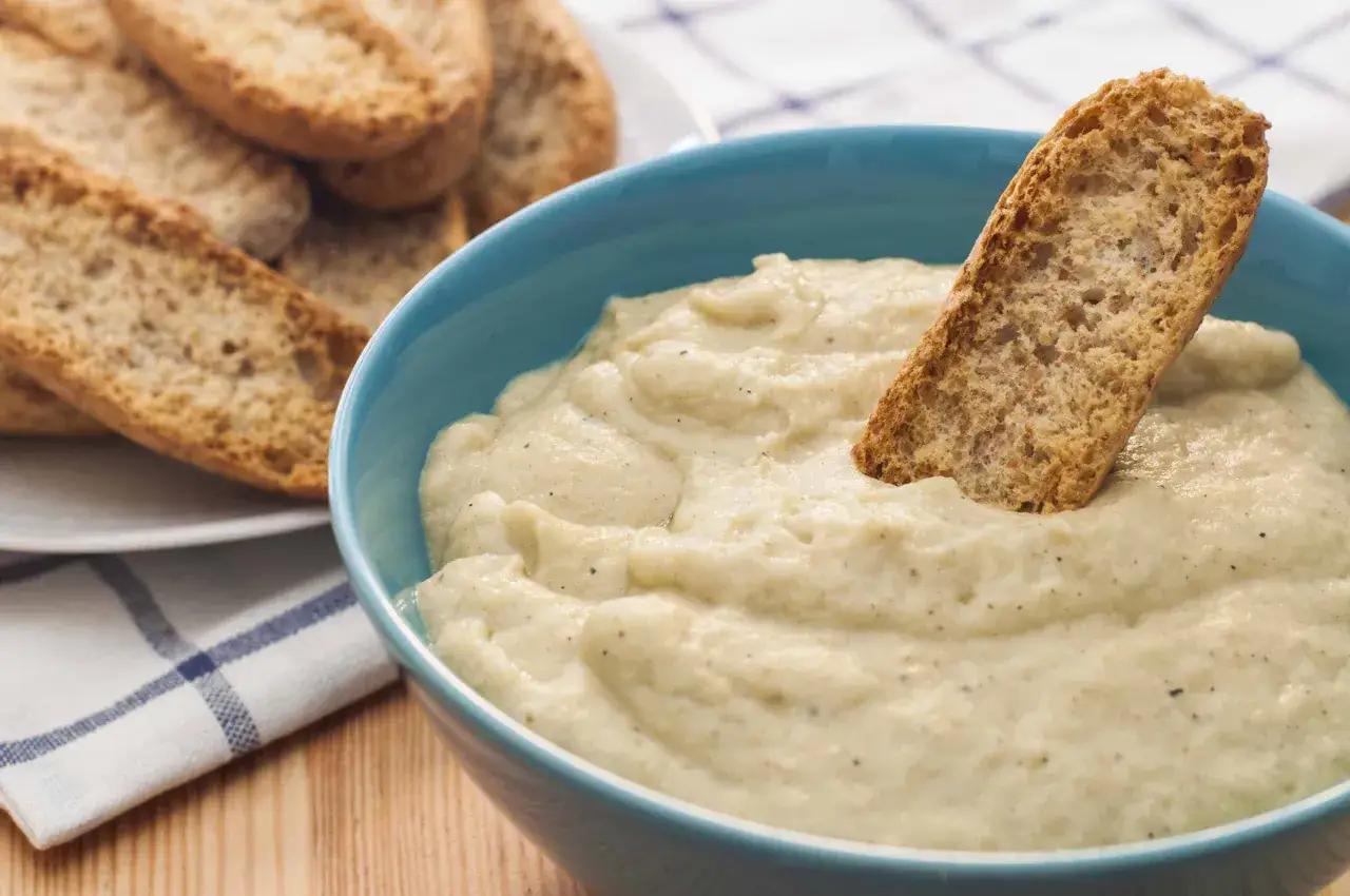Un trozo de pan crujiente se sumerge en un cremoso pure de berenjena. Al fondo, más pan tostado en un plato.