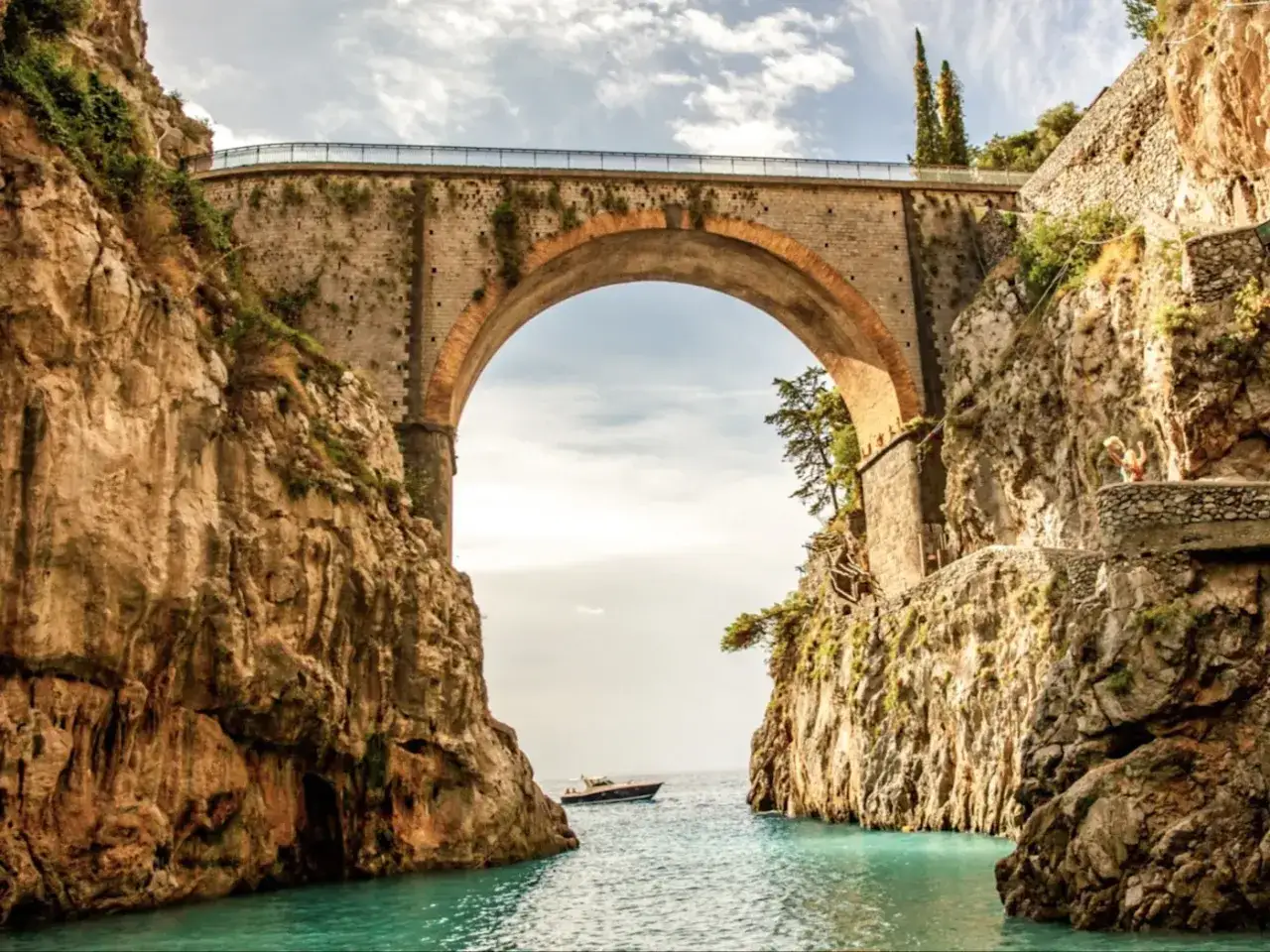Un pont majestueux enjambe une crique aux eaux turquoise, parfait pour des vacances sur la côte amalfitaine. Un bateau navigue paisiblement.