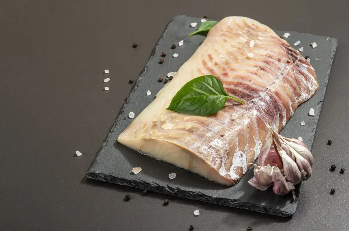 Surowy filet z dorsza czarnego, przyprawiony solą i pieprzem, z czosnkiem i listkami bazylii na czarnej desce.