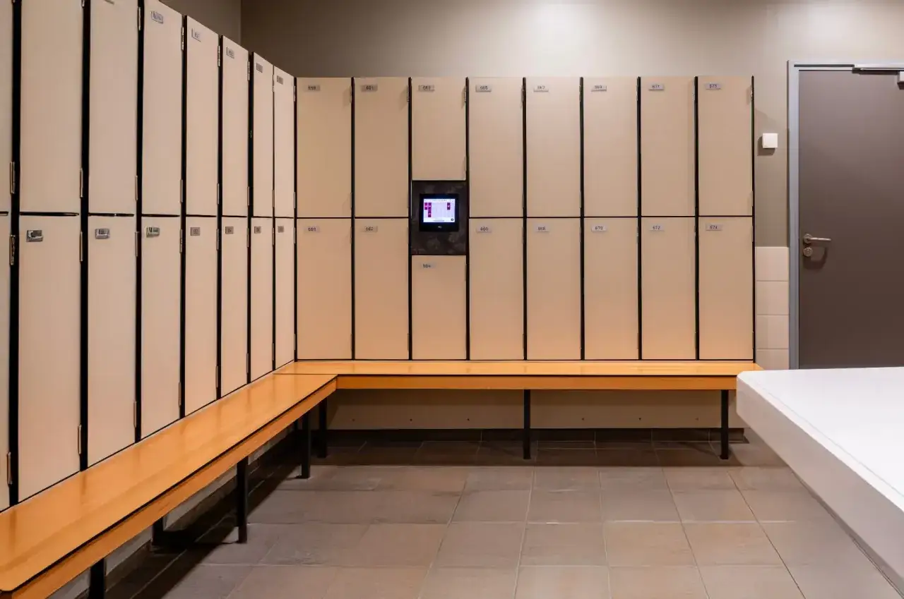 Un vestiaire moderne avec des casiers beiges numérotés et un banc en bois. Un écran tactile est intégré au centre des casiers.