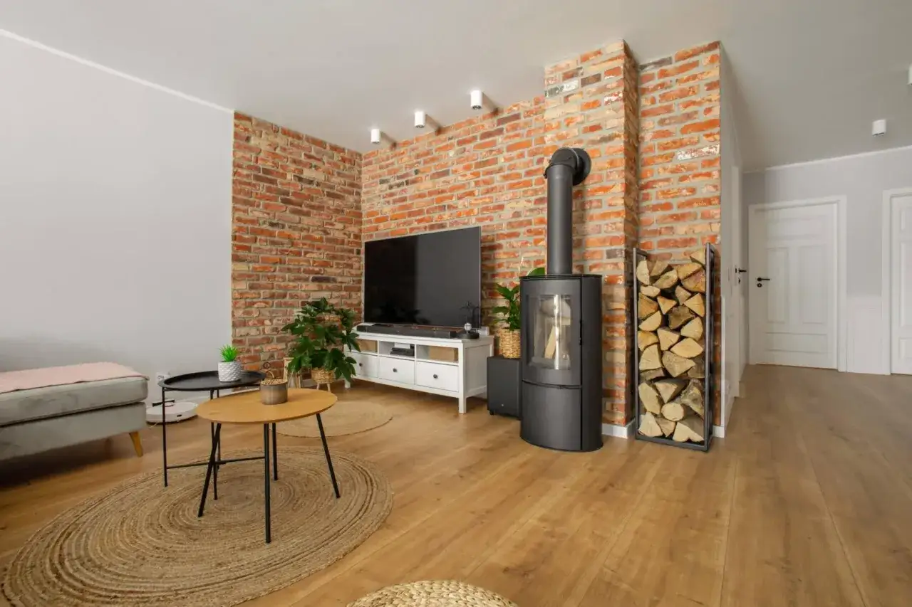 Nowoczesny salon z ceglaną ścianą, czarnym piecykiem i stertą drewna. Kominek z cegły stanowi serce tego przytulnego wnętrza.
