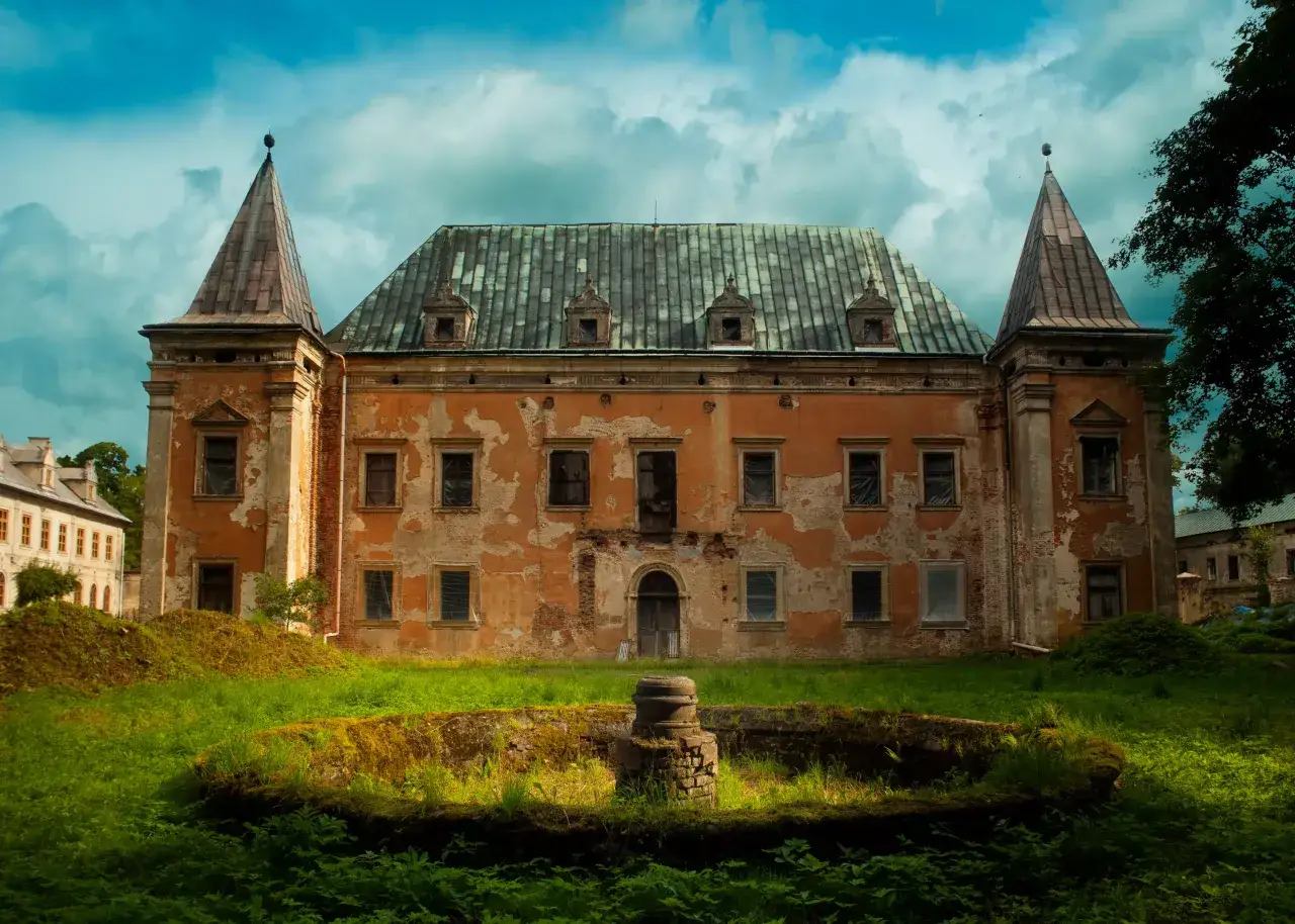 Zrujnowany pałac z pomarańczową fasadą i zielonym dachem, otoczony bujną roślinnością. W oddali widać inny budynek. Kruszyna historii.