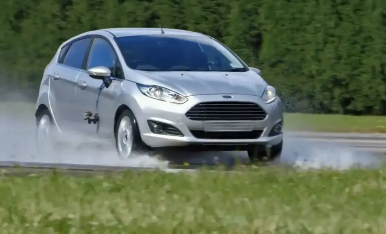 Srebrny Ford Fiesta wpada w aquaplaning na mokrej nawierzchni, tworząc kaskadę wody.