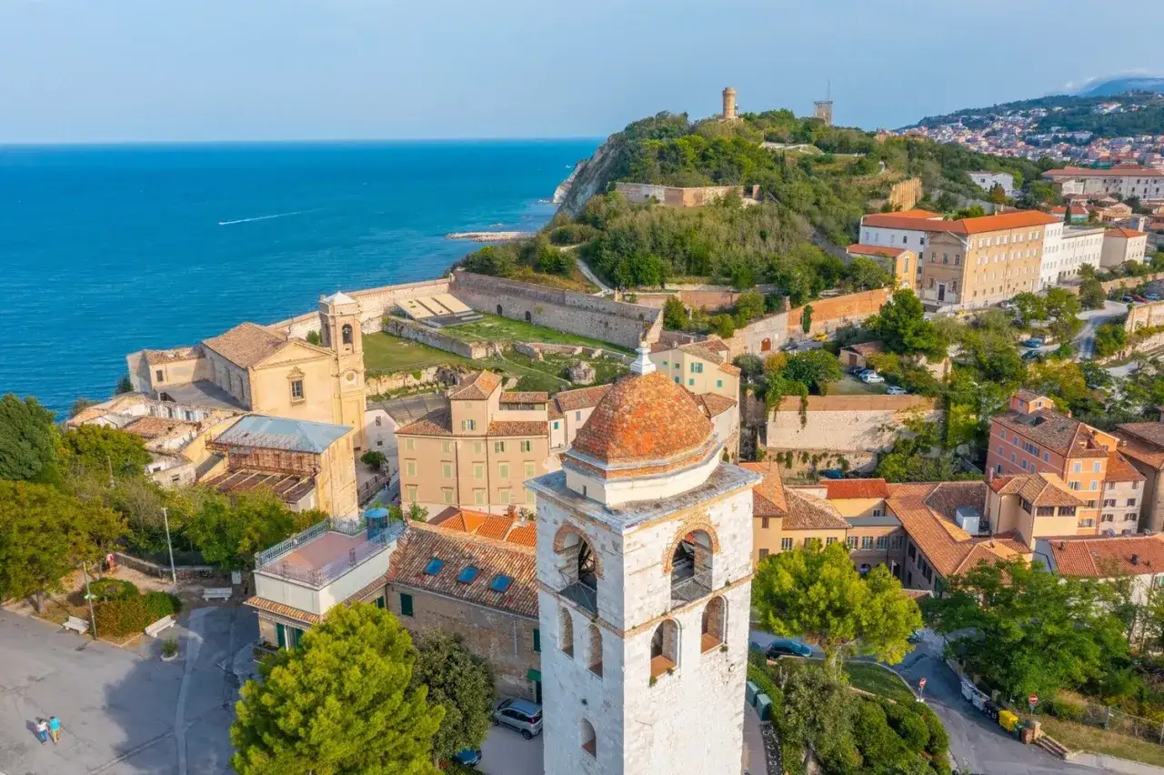 Vista aerea del centro storico di Ancona, con il mare blu, chiese, torri e case con tetti rossi.