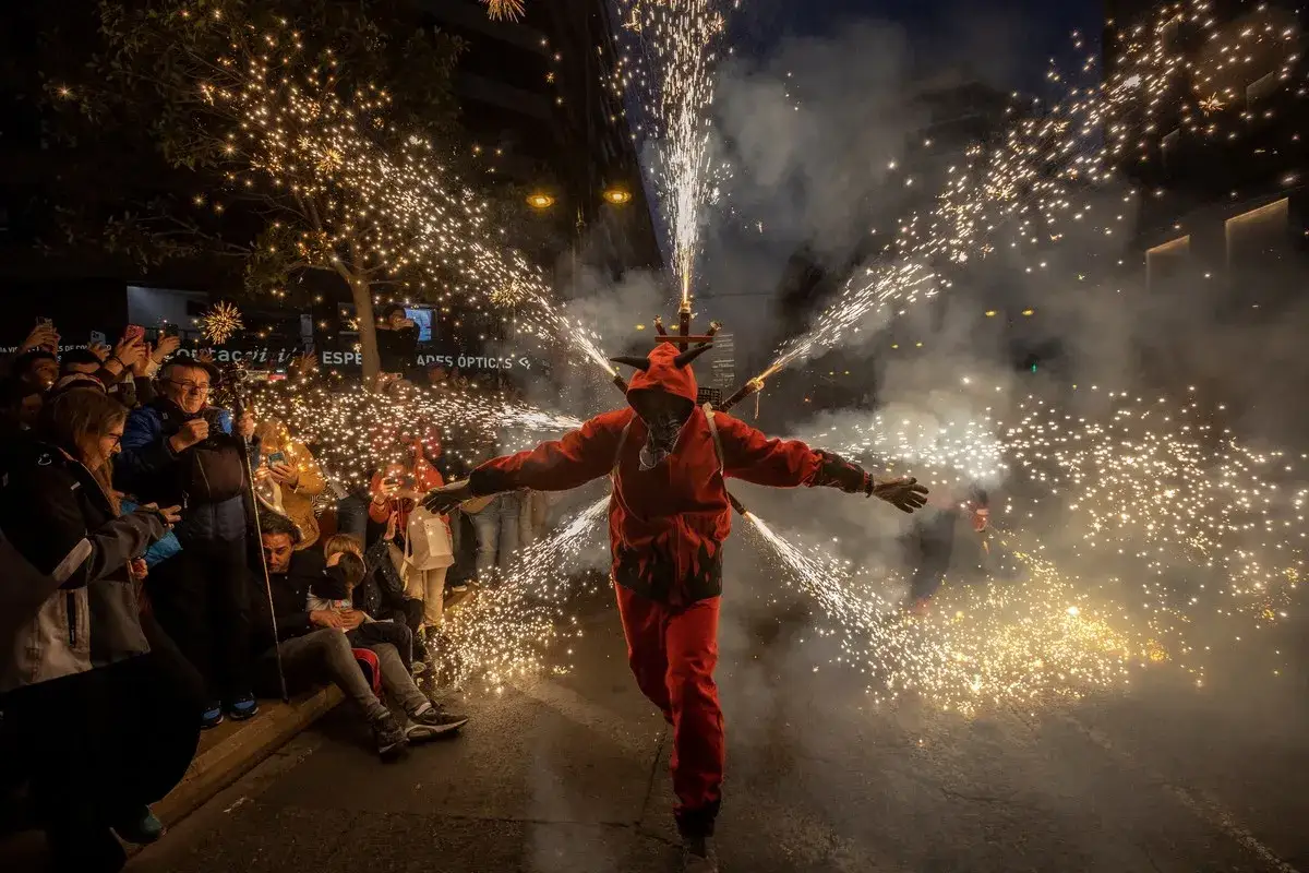 Un diablo con cuernos y traje rojo desata fuegos artificiales en una calle, rodeado de gente expectante. ¡Una escena vibrante que anticipa las fallas 2026!