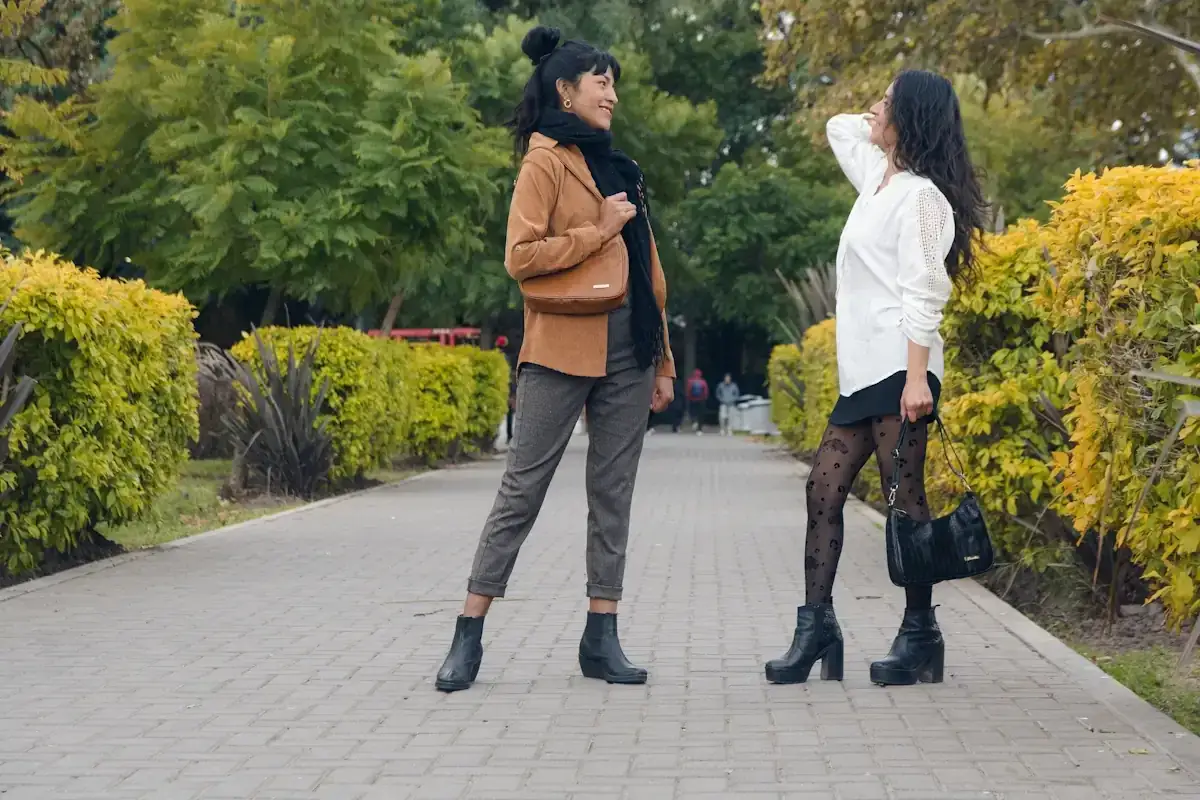 Deux femmes discutent dans un parc. L'une porte une veste marron, un foulard noir et un pantalon gris. L'autre, une chemise blanche, une jupe noire et des collants fantaisie. Idéal pour savoir comment s'habiller quand il fait 15 degrés.