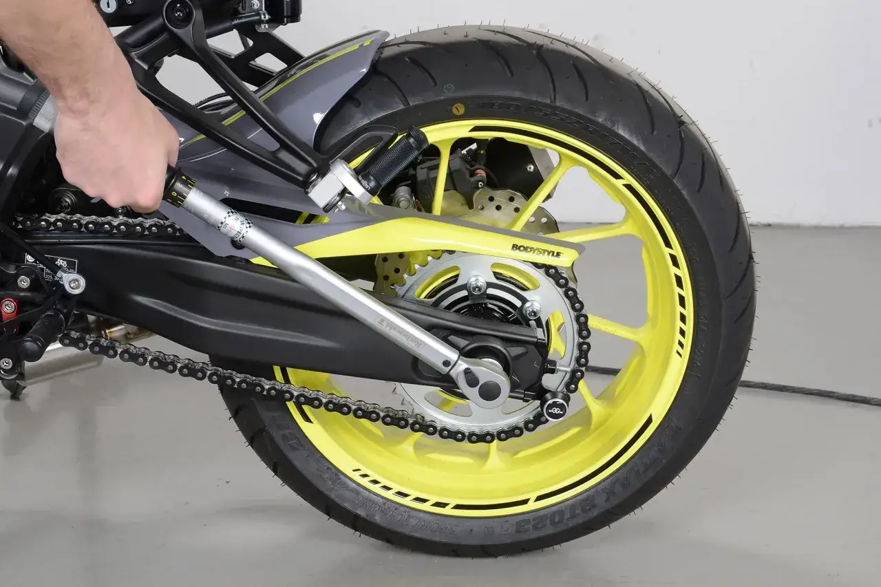 Dłoń trzyma niutonometr, regulując napięcie łańcucha motocykla z żółtą felgą.