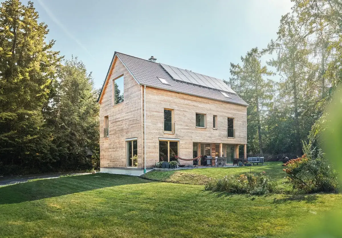 Modernes Architektenhaus Holz mit Solaranlage auf dem Dach, umgeben von Bäumen und einem gepflegten Garten.