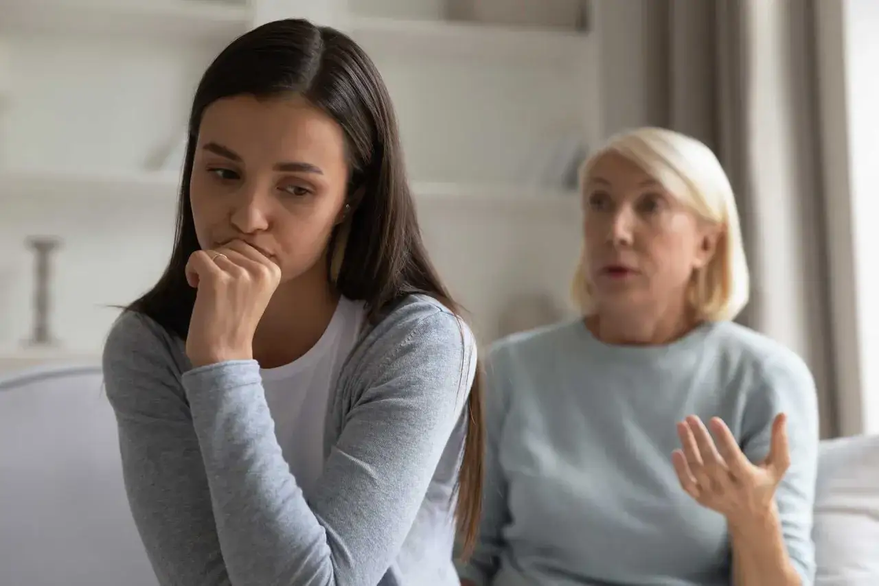 En ung kvinna sitter med handen mot munnen, ser bekymrad ut. Bakom henne gestikulerar en &auml;ldre kvinna, kanske en diskussion om ett problem med vuxen dotter.