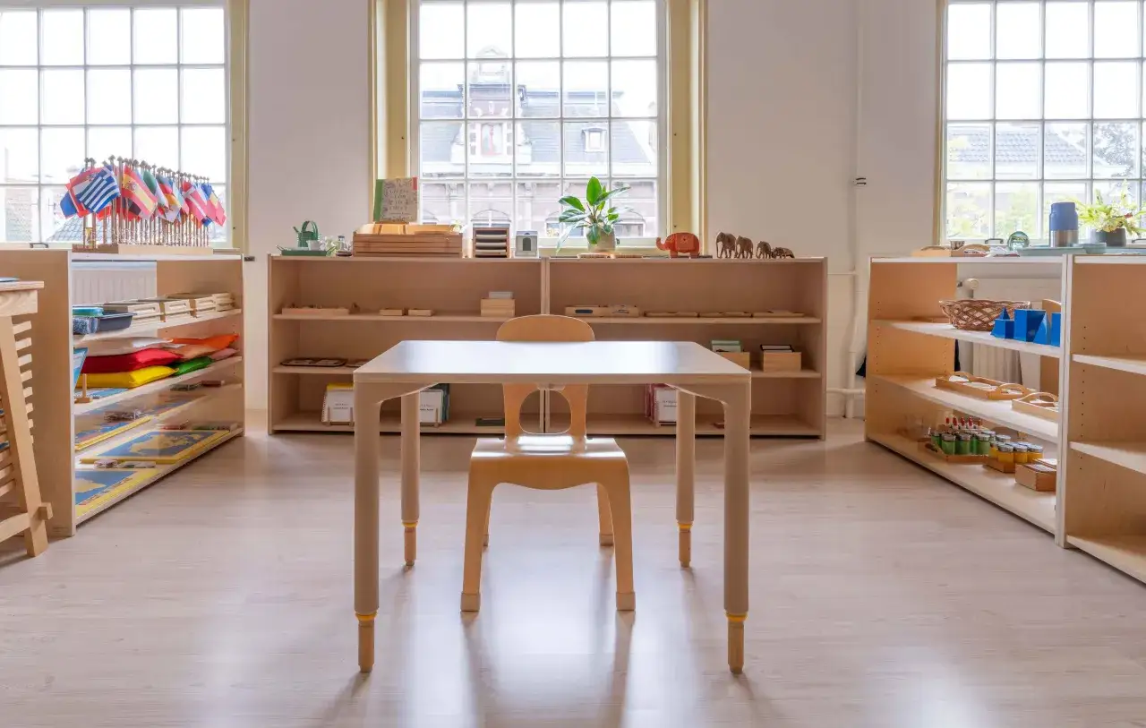 Ein heller Raum mit Regalen voller Montessori-Materialien. Ein kleiner Tisch mit einem Stuhl bildet die Sitzordnung.