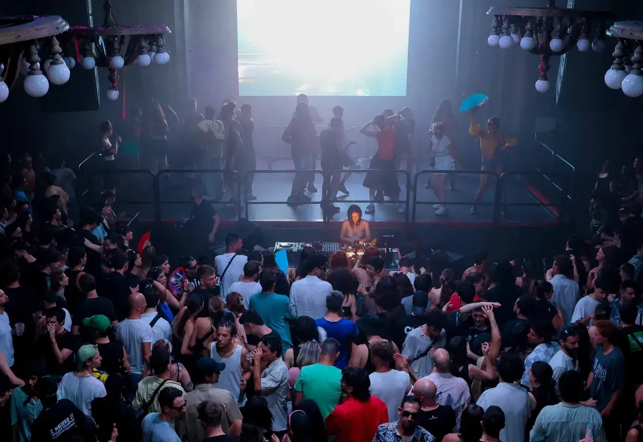 Una folla balla al Nitsa Club, illuminata da un DJ set e da un grande schermo. L'atmosfera &egrave; vibrante e piena di energia.