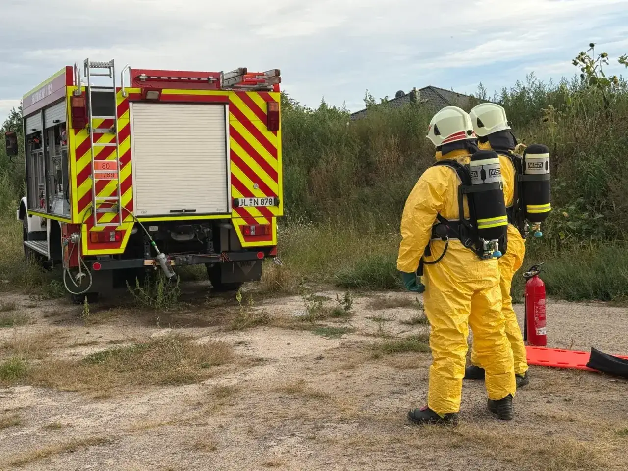 Zwei Einsatzkr&auml;fte der Feuerwehr in gelben Chemikalienschutzanz&uuml;gen stehen bereit. Ein L&ouml;schfahrzeug steht im Hintergrund.