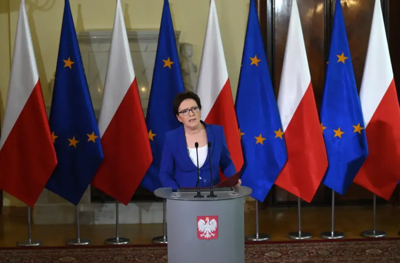 Ewa Kopacz, premier, przemawia na tle flag Polski i Unii Europejskiej.