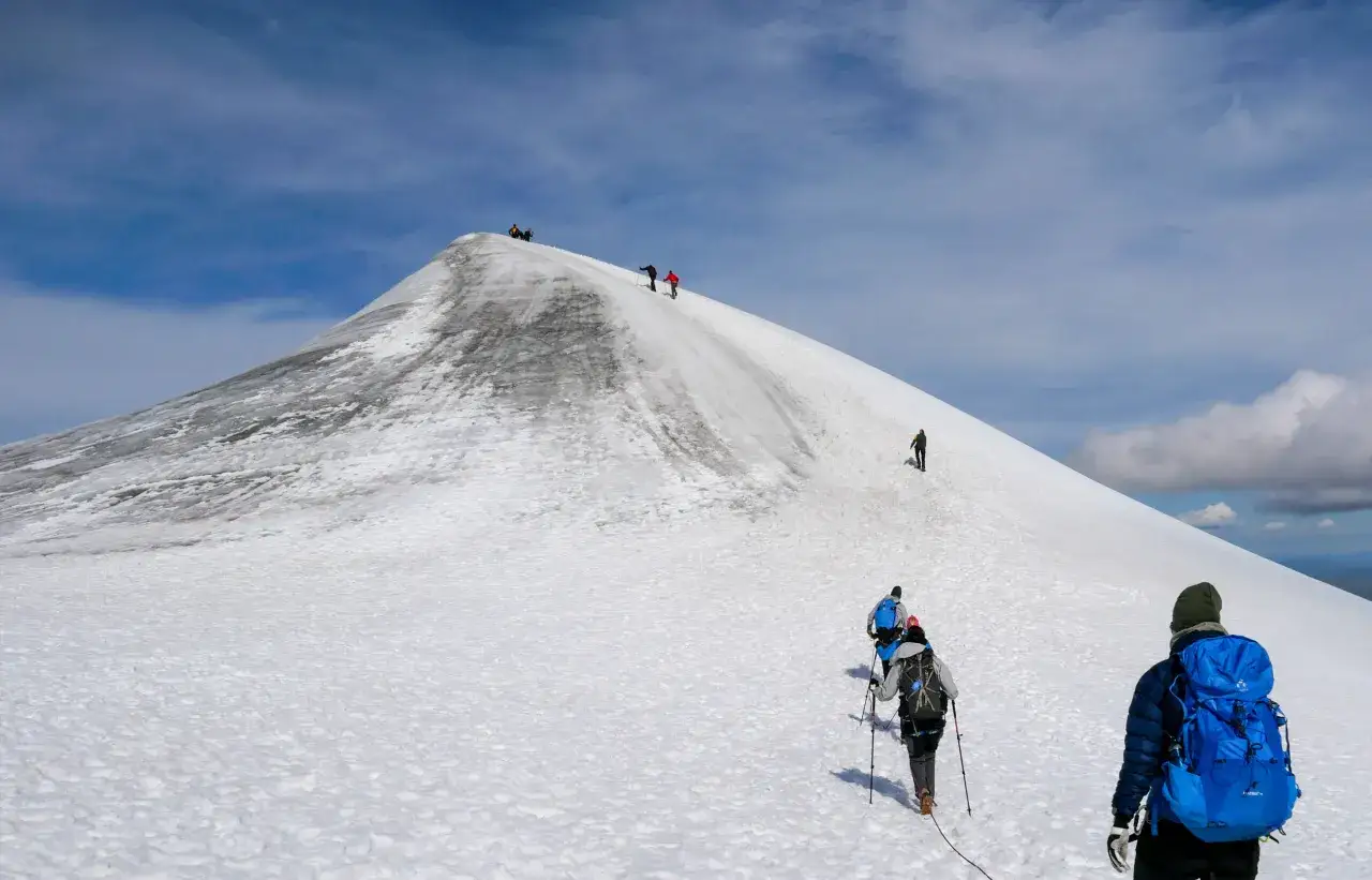 Vandrare kl&auml;ttrar uppf&ouml;r en sn&ouml;ig sluttning p&aring; Kebnekaise, med en bl&aring; himmel och moln ovanf&ouml;r. Ingen lavin syns, men terr&auml;ngen antyder dess potential.