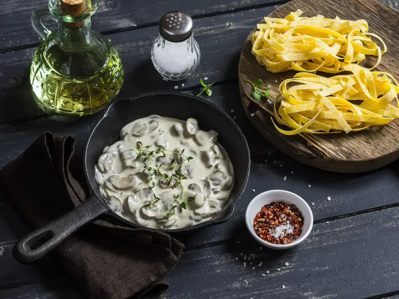 Kremowy sos pieczarkowy w patelni, obok świeżego makaronu tagliatelle, butelki oliwy i przypraw. Idealny do przygotowania pysznego dania.