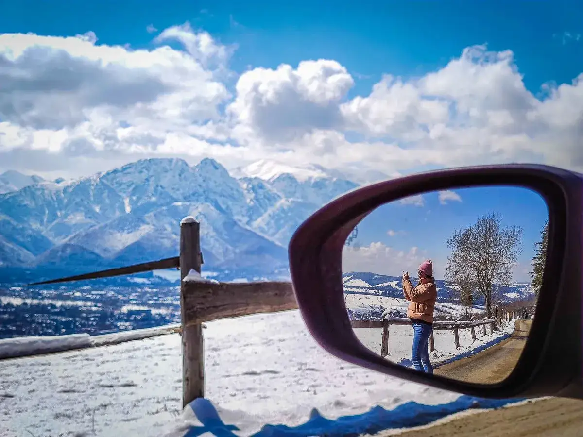 Widok na zaśnieżone Tatry z bocznego lusterka samochodu. Dziewczyna robi zdjęcie na zakopiance.