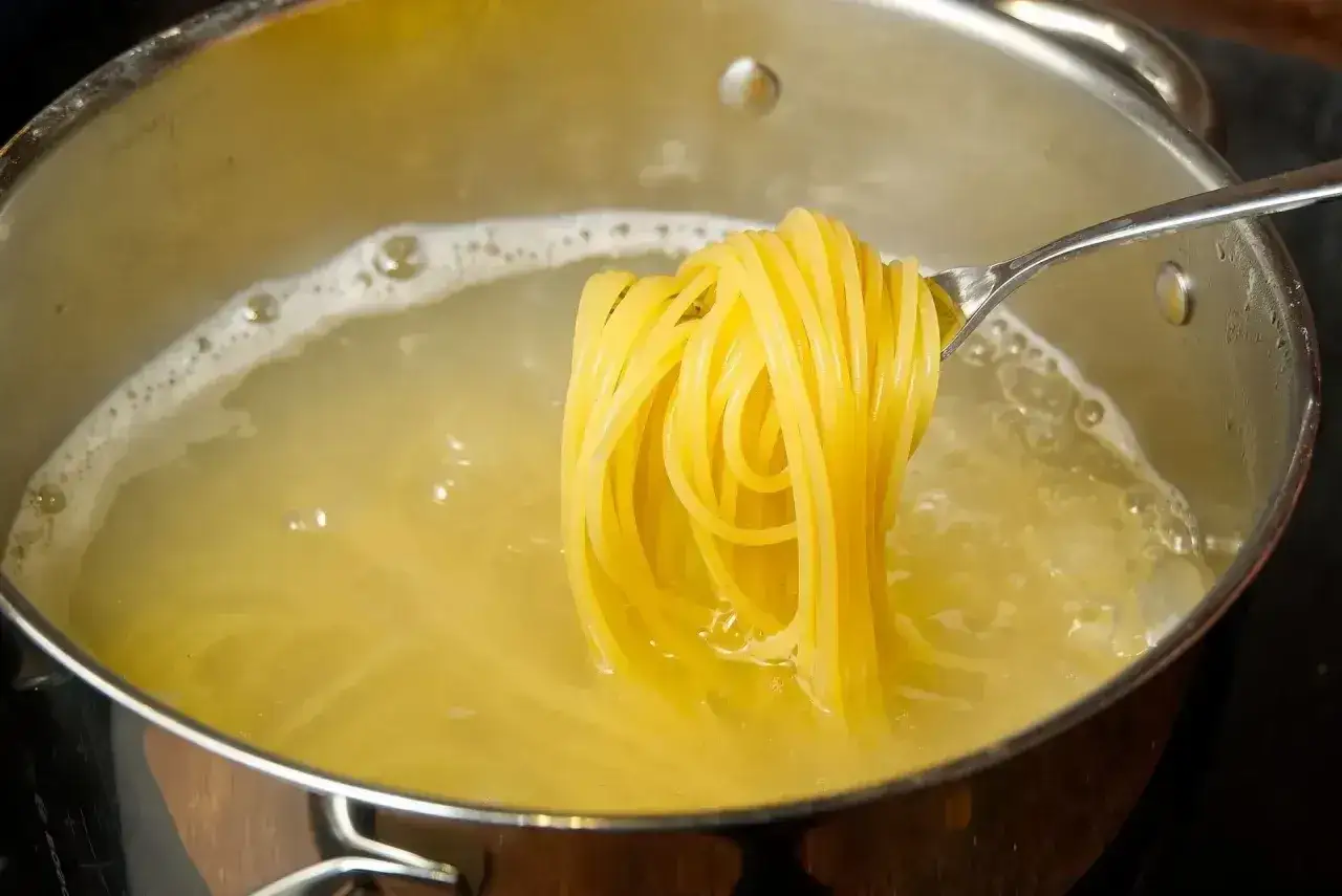 Z widelca zwisają ugotowane spaghetti, bogate w węglowodany, unosząc się nad bulgoczącą wodą w garnku.