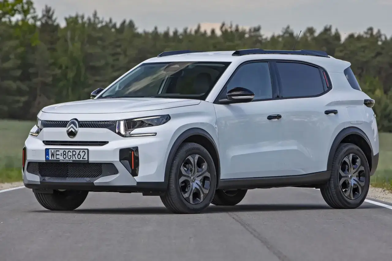 Biały, **mały SUV** Citroën C3 Aircross z czarnymi relingami dachowymi i przyciemnianymi szybami jedzie po asfaltowej drodze.