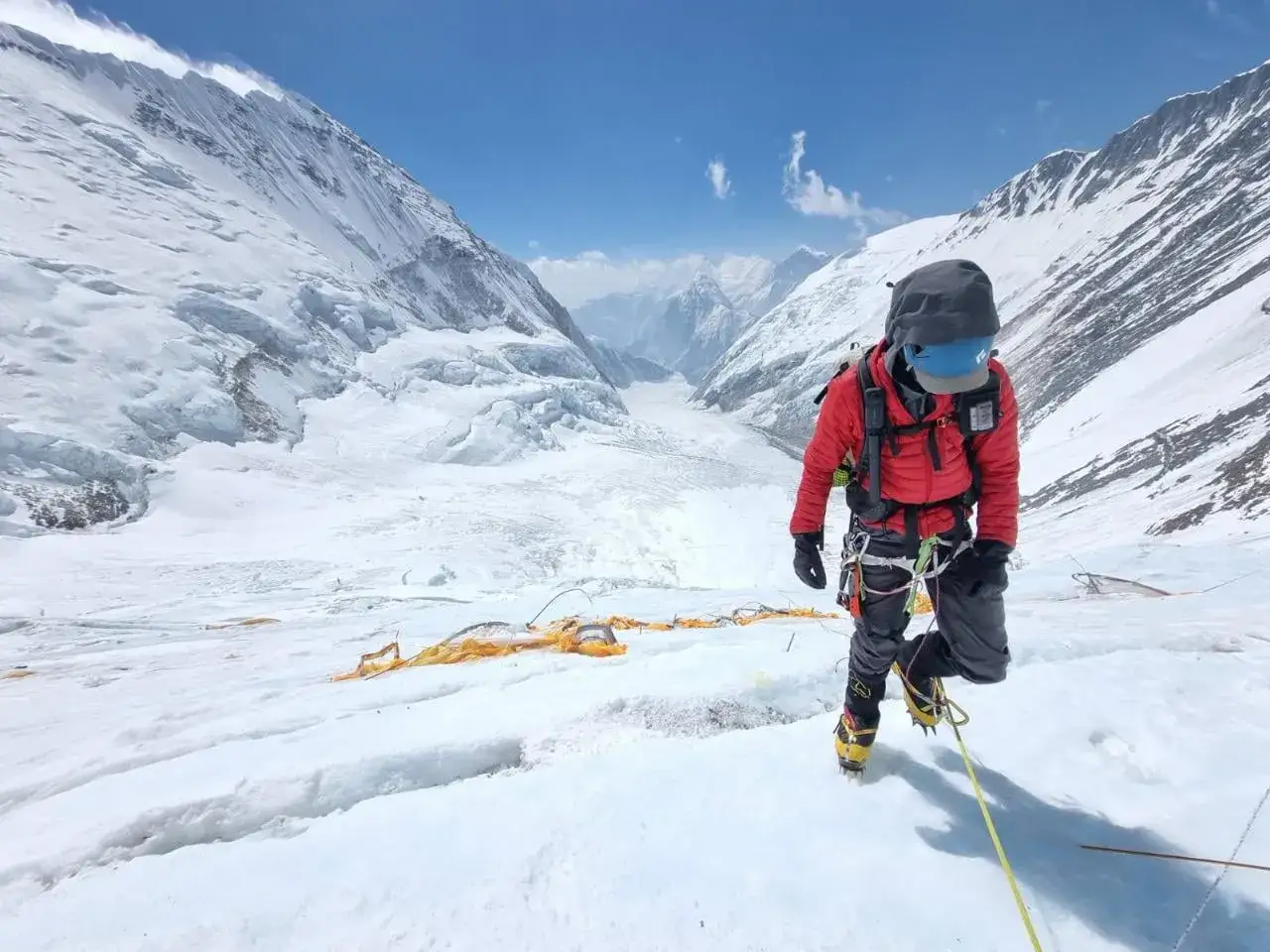 Alpinista w czerwonej kurtce schodzi po ośnieżonym zboczu, korzystając z poręcz&oacute;wek w Himalajach. W tle rozciąga się lodowiec i ośnieżone szczyty.