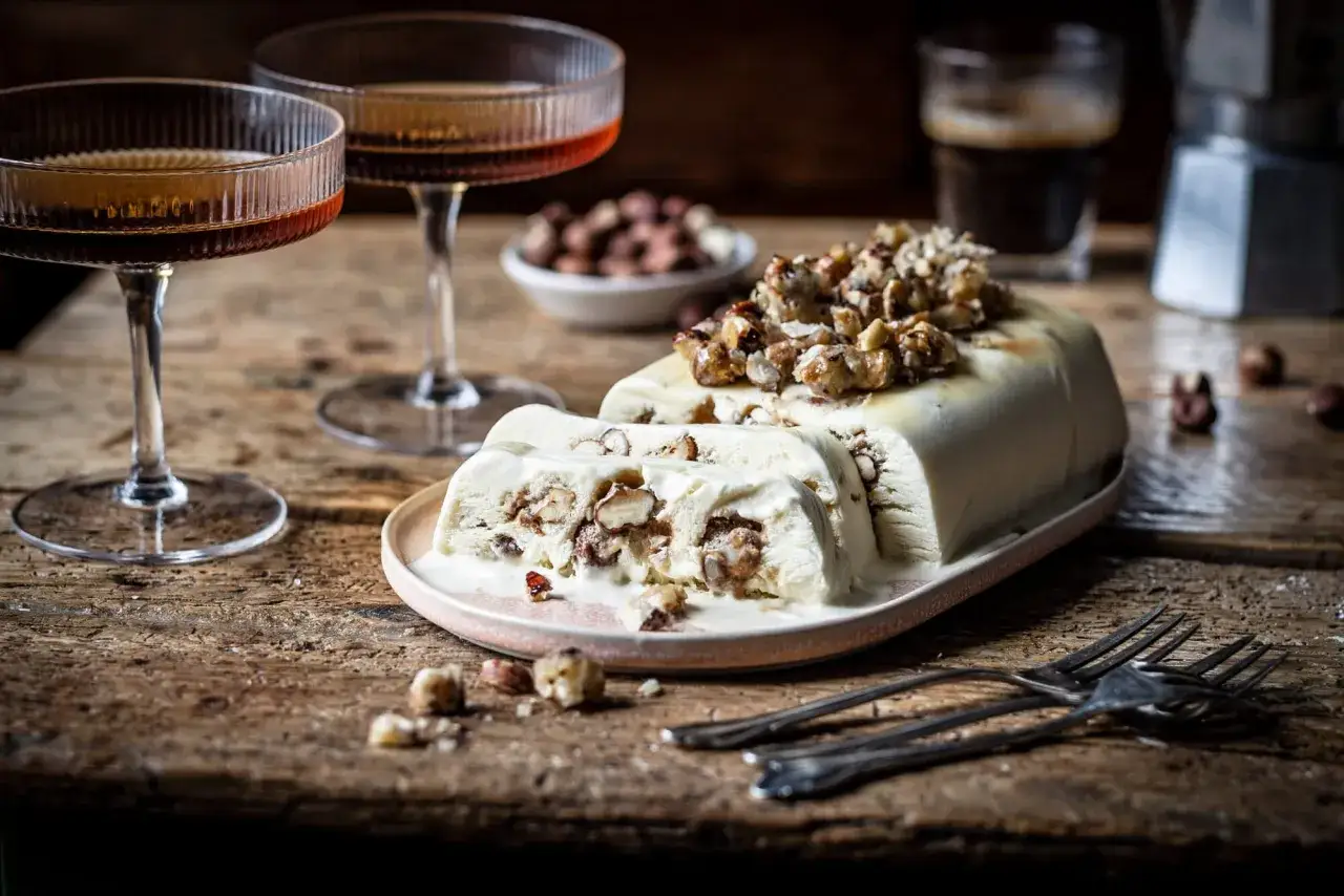Cremiges Nougat-Eis mit N&uuml;ssen, serviert auf einem Teller mit zwei Gl&auml;sern Lik&ouml;r und einer Mokkakanne im Hintergrund.