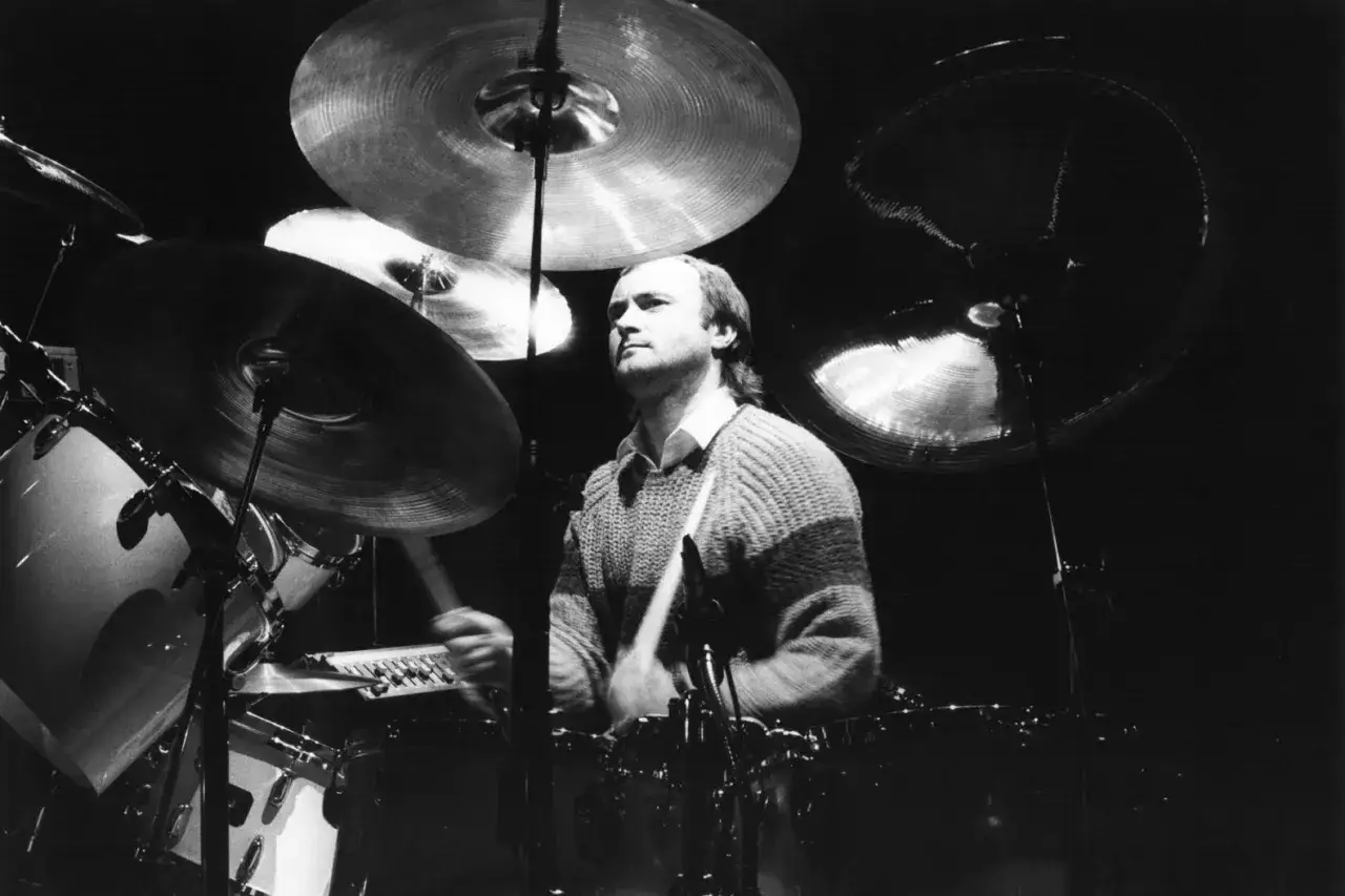 Phil Collins w akcji przy perkusji, jego dłonie z pałeczkami unoszą się nad bębnami, tworząc rytm.