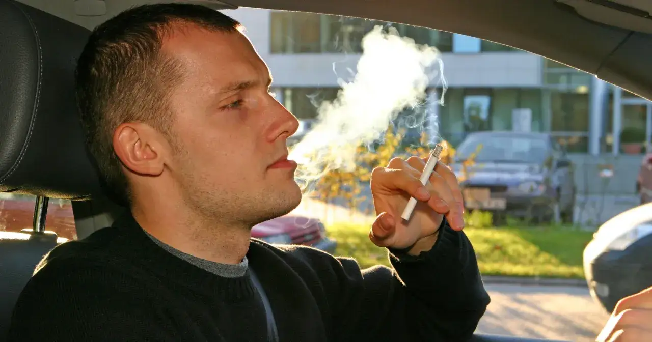 Mężczyzna pali papierosa w samochodzie, a dym unosi się w powietrzu. Zastanawiasz się, jak pozbyć się zapachu papierosów z auta?