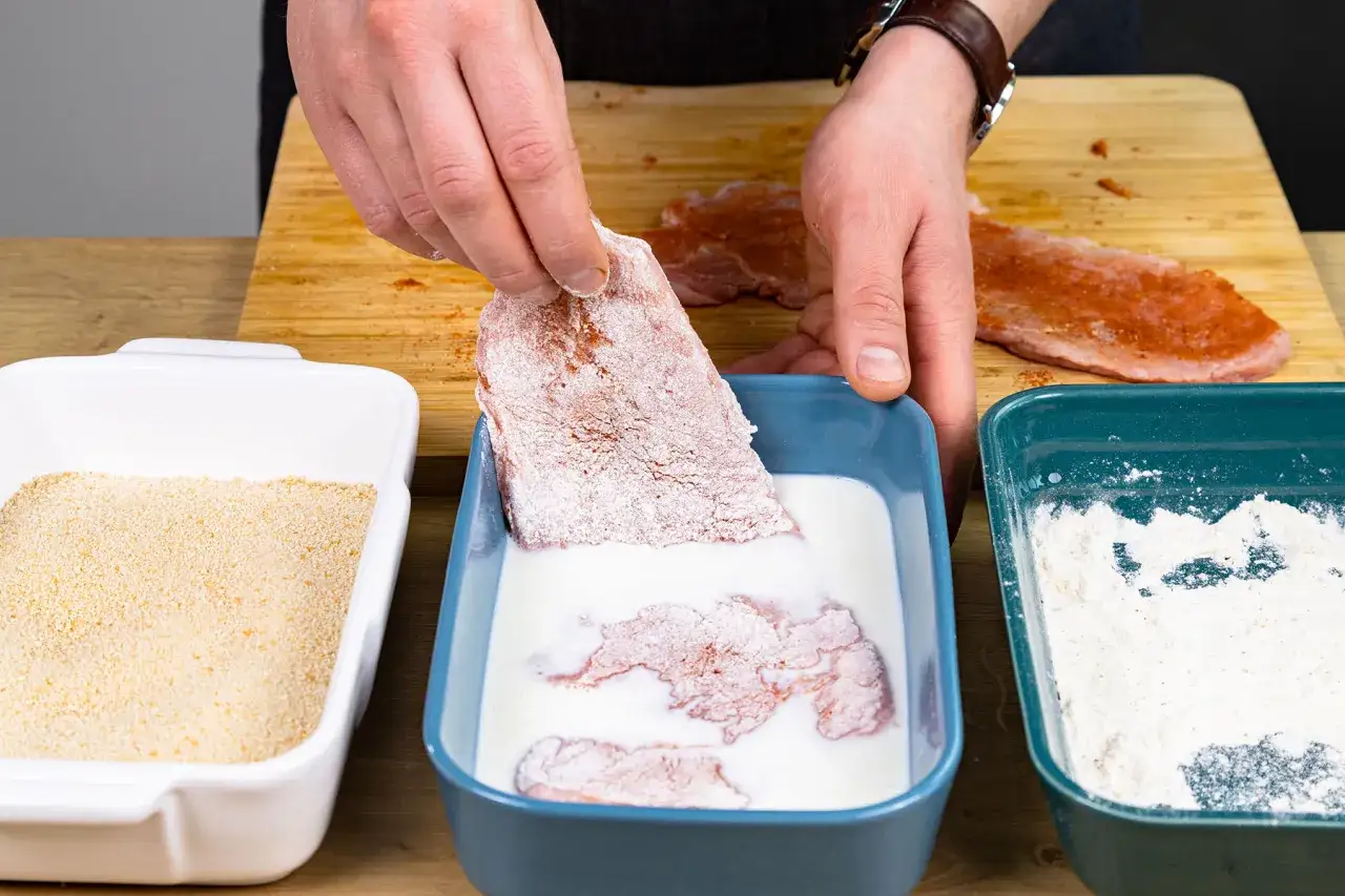 Fleisch wird in eine weiße Flüssigkeit getaucht, um es für das Panieren ohne Ei vorzubereiten. Daneben liegen Paniermehl und Mehl.