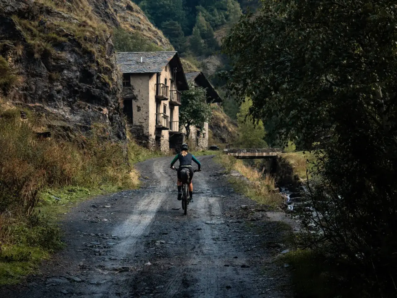 Persona pedaleando por un camino de grava hacia unas casas de piedra en la monta&ntilde;a. Una aventura de viajar en bicicleta.