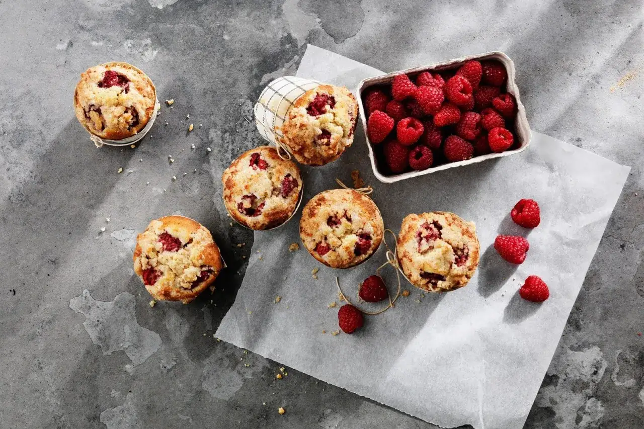 Sju mumsiga cupcakes med fyllning av hallon ligger utspridda på ett grått underlag, bredvid en korg full med färska hallon.