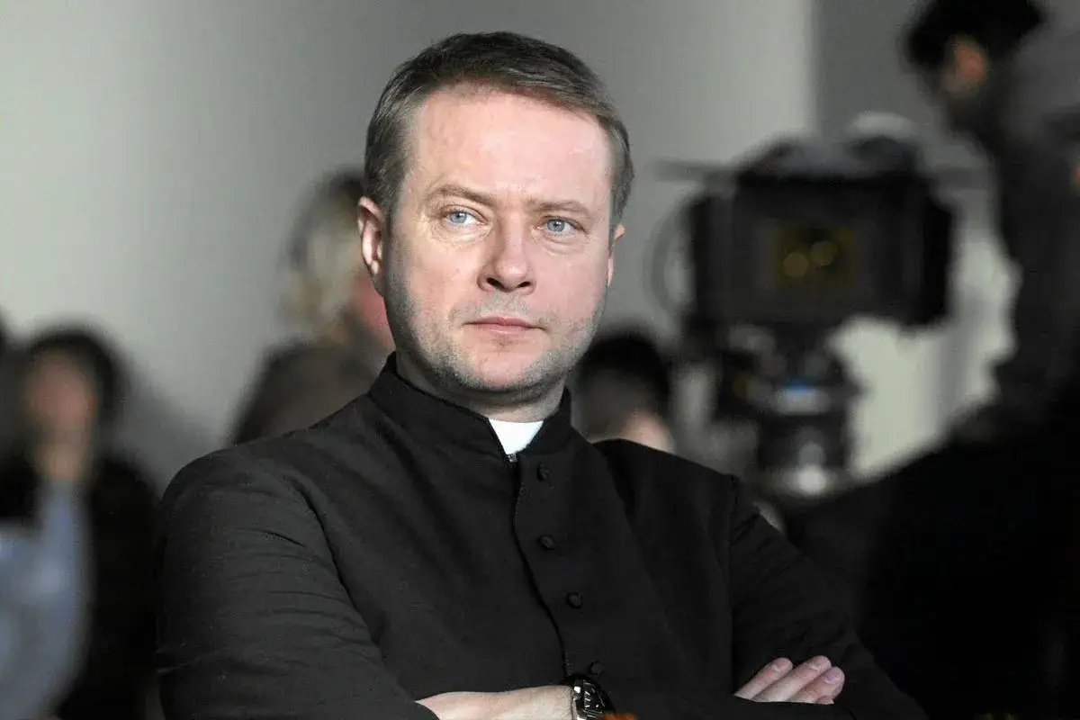 Artur Żmijewski w roli księdza na planie filmowym. W tle widać ekipę filmową i sprzęt.