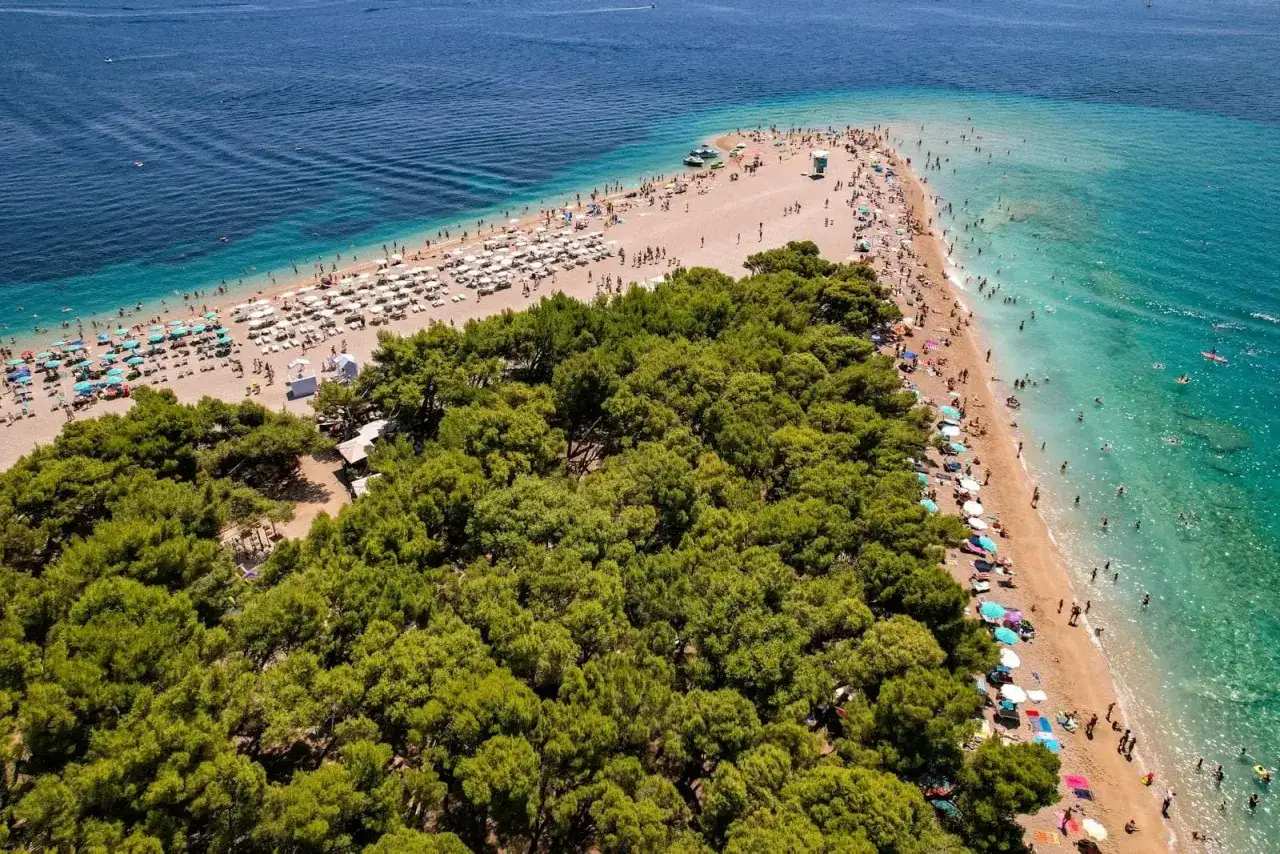 En solig strand med många parasoller och badande människor vid Zlatni Rat, en berömd udde på ön Brač i Kroatien.
