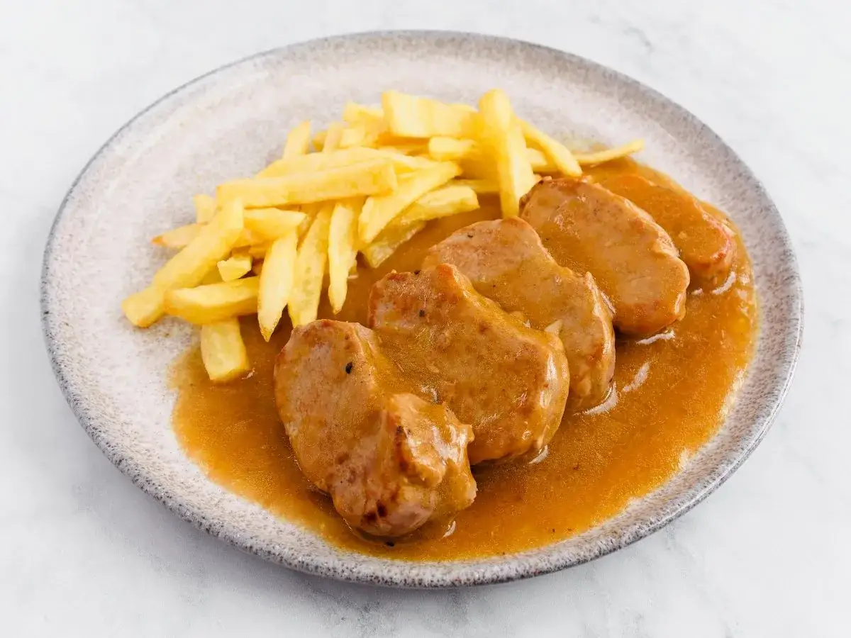 Cuatro trozos de solomillo al whisky en una salsa dorada, acompa&ntilde;ados de patatas fritas crujientes en un plato.