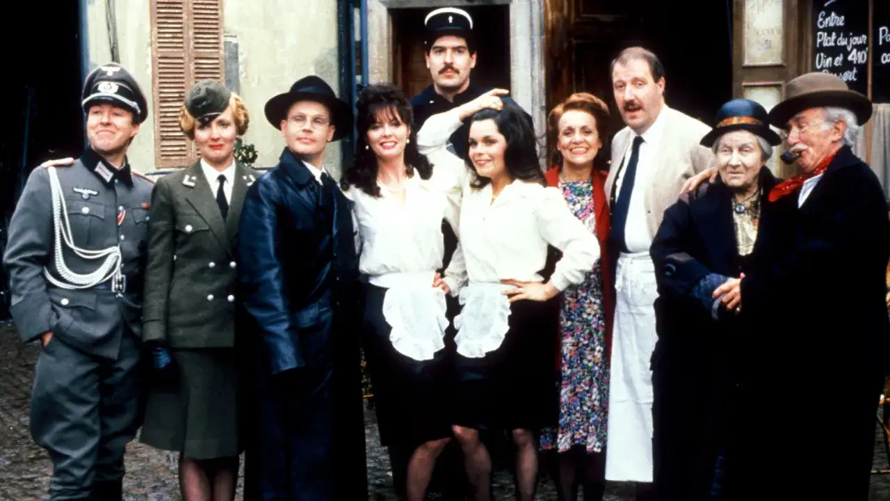 Allo allo! Grupa postaci w strojach z serialu, w tym żołnierze, kelnerki i starsi ludzie, pozuje do zdjęcia.