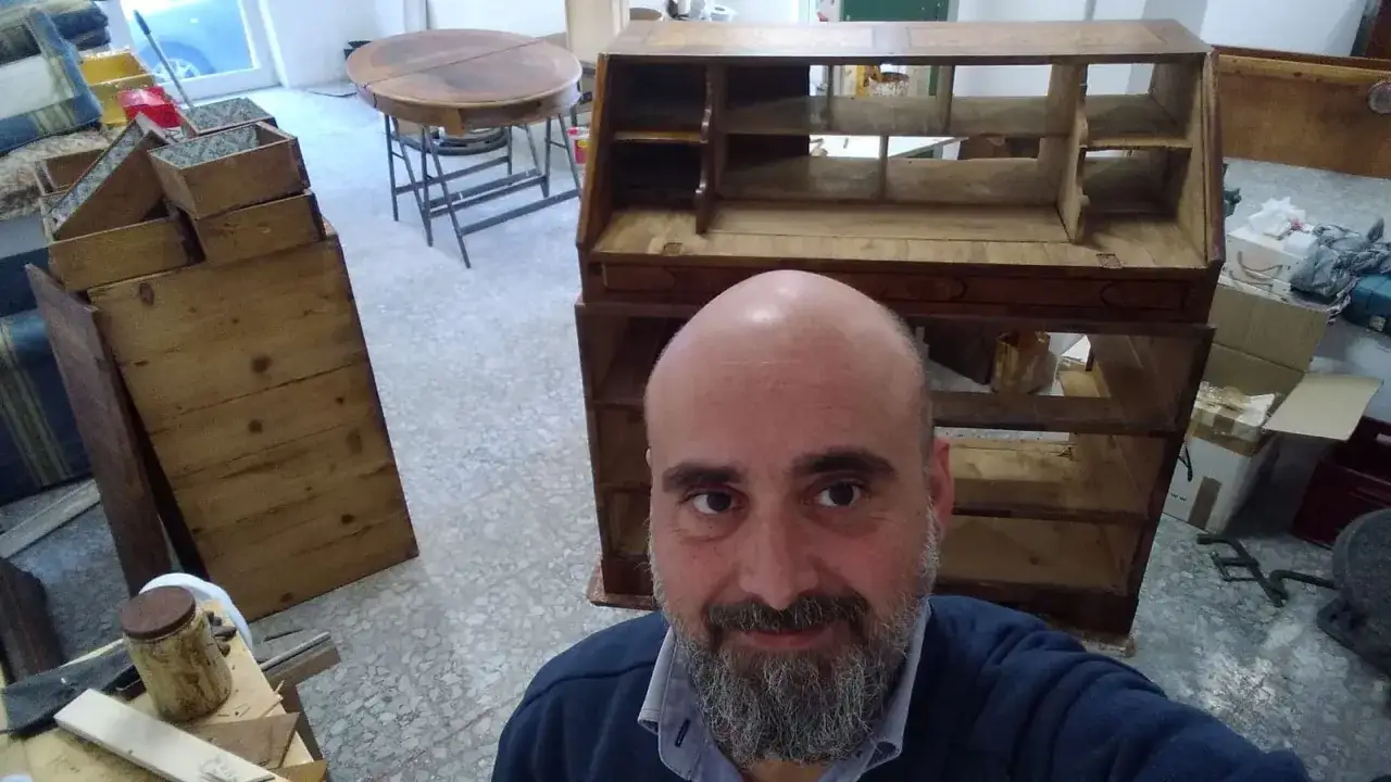 Un falegname mostra come si lavora il legno, con mobili antichi e attrezzi da lavoro in un laboratorio.