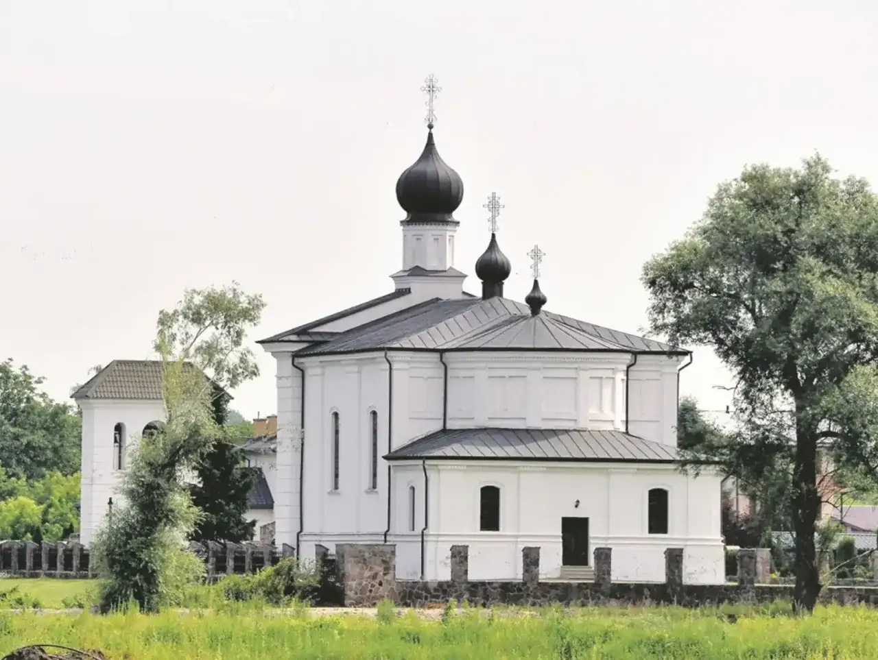 Biała cerkiew z czarnymi kopułami w Terespolu, otoczona zielenią i kamiennym murem.