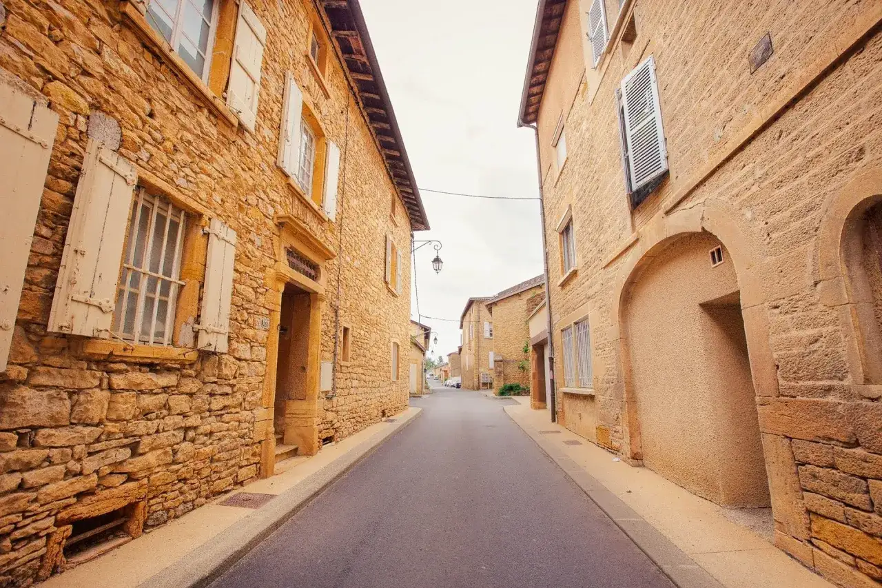 Rue pavée bordée de maisons en **pierre dorée**, aux volets clos, sous un ciel nuageux. Ambiance de village authentique.
