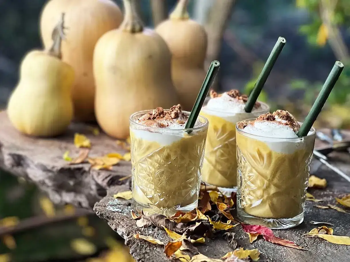 Tre glas med kr&auml;mig pumpa- och honungsdrink, toppad med gr&auml;dde och n&ouml;tter. Pumpan i bakgrunden skvallrar om h&ouml;st.