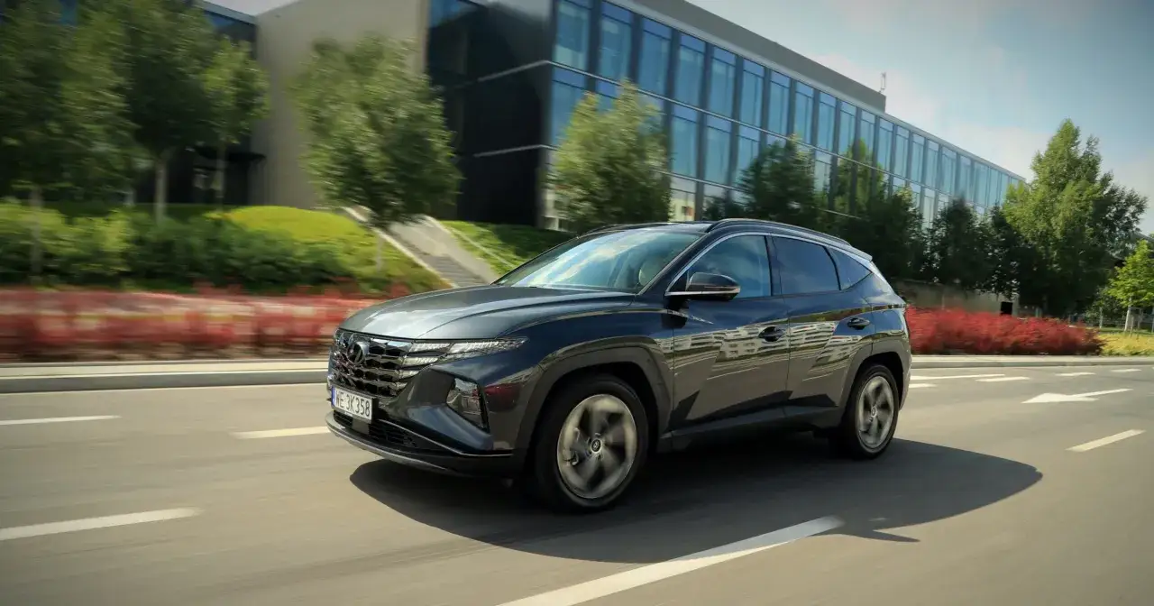Ciemnoszary Hyundai Tucson 4 jedzie drogą obok nowoczesnego budynku i zieleni.